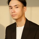 A professional portrait of a young man wearing a black suit indoors.